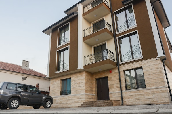 A multi-story residential building with balconies, a car parked in front, and a door with a red handle.