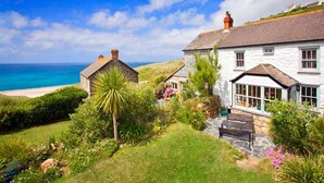 A coastal house with a stone exterior, a garden with various plants, and a view of the sea. - Petra, Beach Cottage, Great Sea Views, Garden (Sennen)