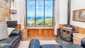 A cozy living room with a stone wall, a fireplace, a large window with a view of the beach, and a television. - Girl Annie, Beach Cottage, Great Sea-Views, Garden (Sennen)