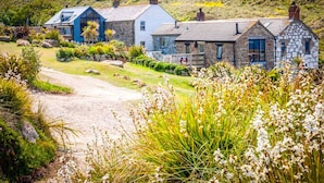 A coastal property with stone and blue buildings, a gravel path, and surrounding vegetation. - Girl Annie, Beach Cottage, Great Sea-Views, Garden (Sennen)