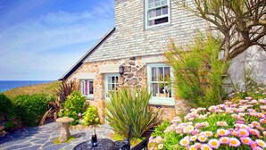 Una casa de piedra con un patio de piedra, una mesa con sillas y un cantero con flores rosas.