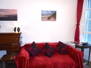 A bedroom with a red bedspread, a wooden dresser, a small table with a red object, a picture on the wall, and a window with red curtains.