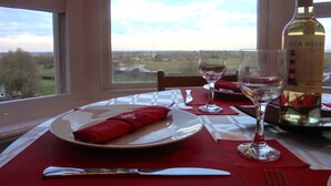 Taula de menjador preparada amb plat, gots de vi i ampolla, amb vista a un paisatge a través d'una finestra.