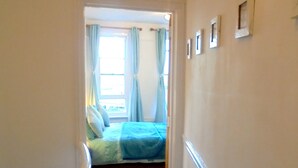 Un passadís estret que condueix a un dormitori amb llit, una finestra amb cortines i un sòl amb moqueta.