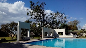 A swimming pool with a stone deck, a white structure with a glass door, and a large tree in the background. - Skyview Lodge At The Barn (Lusaka)