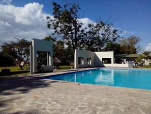 A swimming pool with a stone deck, a white structure with a glass door, and a large tree in the background. - Skyview Lodge At The Barn (Lusaka)