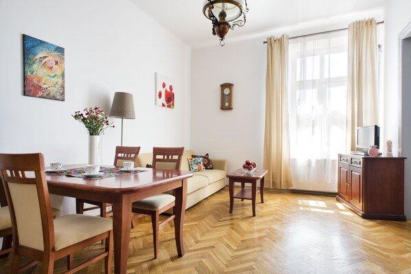A living room with a wooden dining table, chairs, a sofa, a small side table, a lamp, a clock, and a window with curtains.