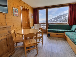 A wooden interior with a dining table, chairs, a sofa, and a view of a snowy mountain landscape through large windows.