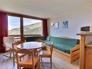 A room with a wooden table, chairs, a sofa, a television on a wooden stand, and a large window with a view of a mountainous landscape.