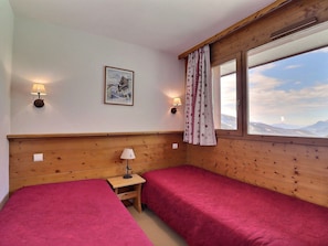 A room with two beds, a small table with a lamp, a painting on the wall, and a window with a view of mountains.