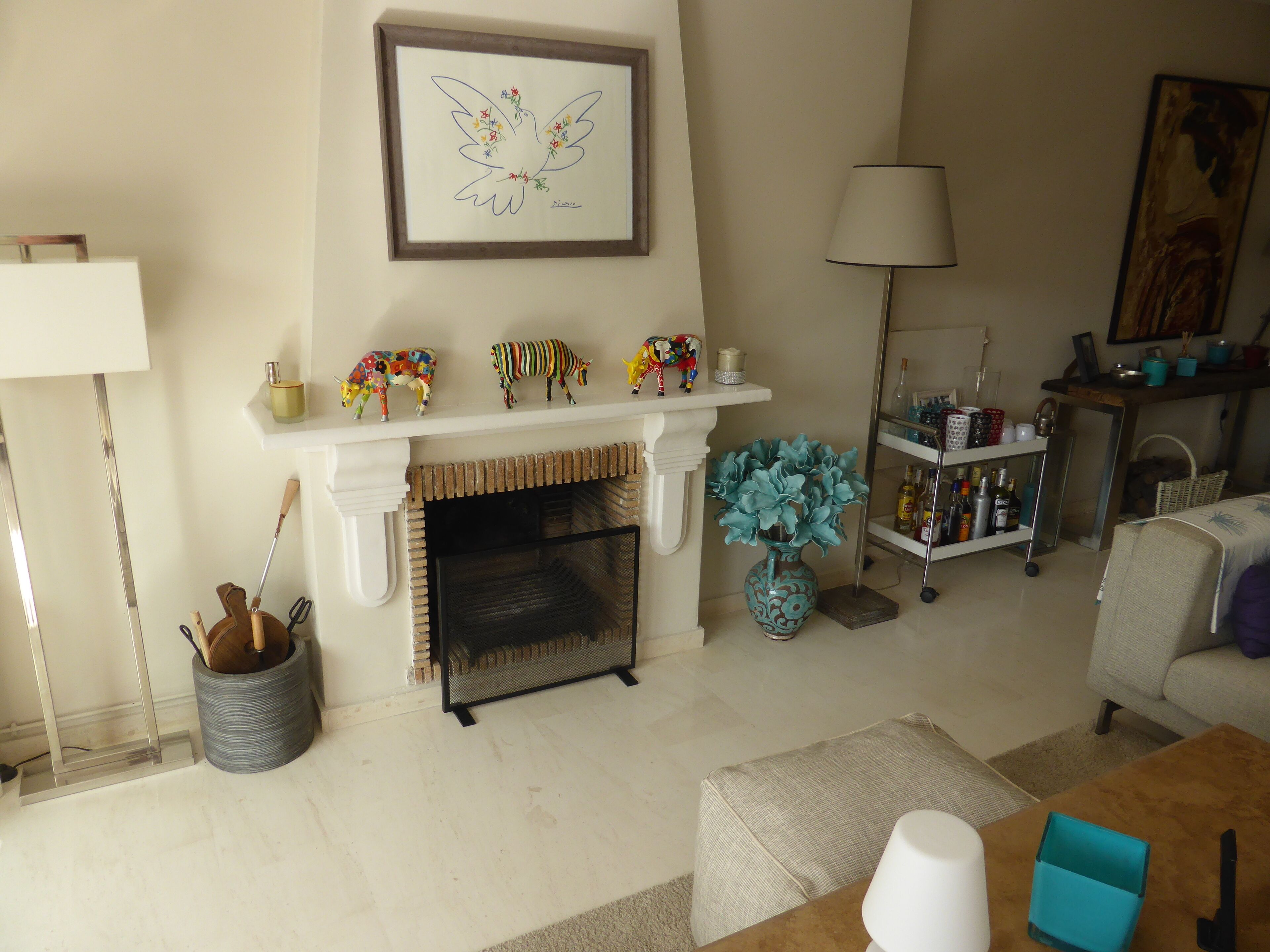 Una sala de estar con chimenea, una repisa decorada con figuritas, un jarrón con flores y una lámpara.