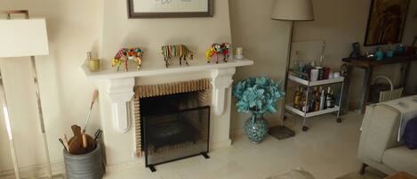 Una sala de estar con chimenea, una repisa decorada con figuritas, un jarrón con flores y una lámpara.