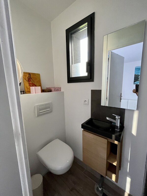 Et lille, moderne badeværelse med et hvidt toilet, en sort vask og et spejl over vasken. Der er en lille hylde med et maleri og en kasse med teksten 'Laura'.