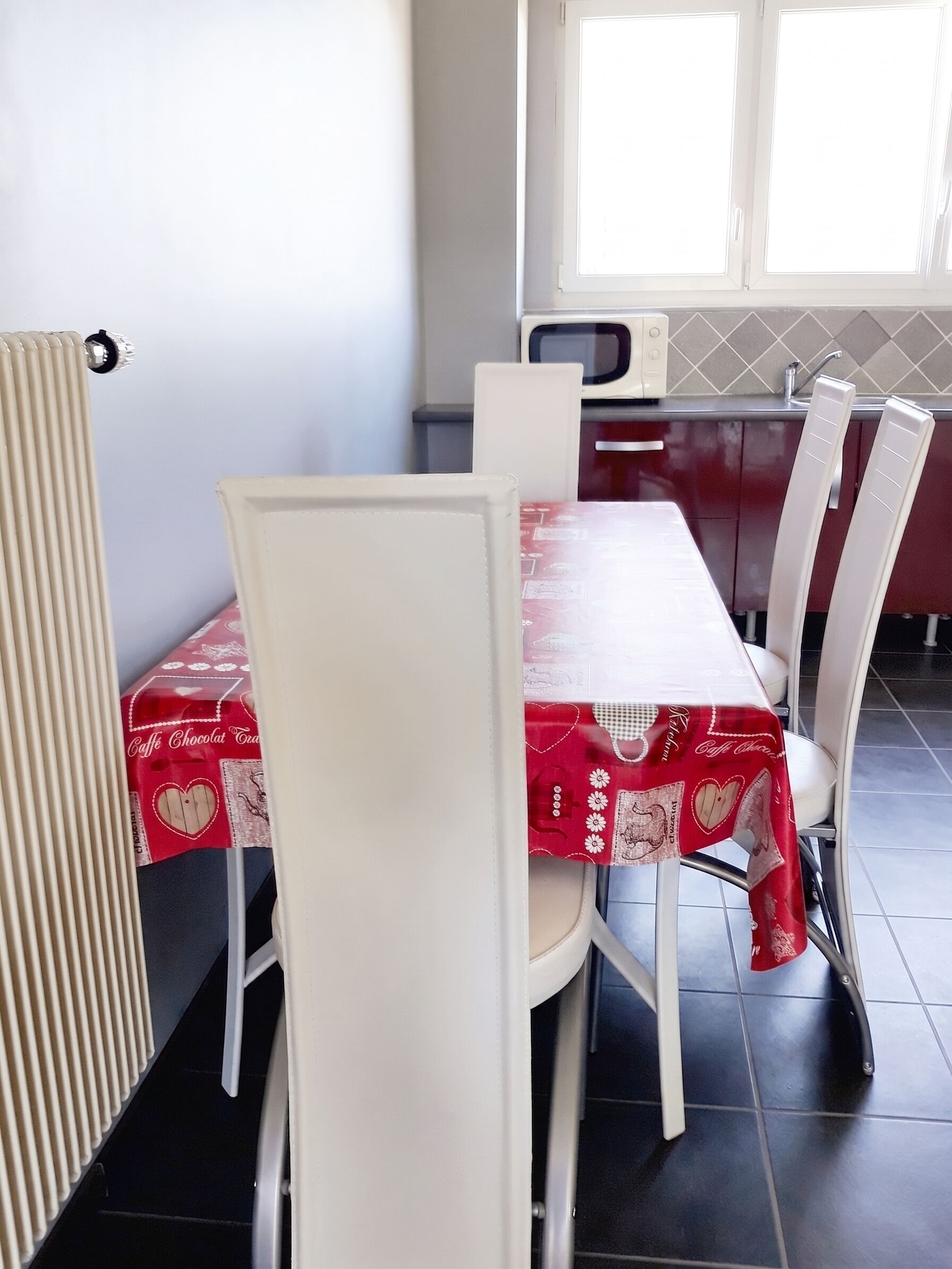 Uma área de refeições com uma mesa coberta por um tecido vermelho, cadeiras brancas, um radiador e uma cozinha ao fundo.
