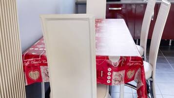 Uma área de refeições com uma mesa coberta por um tecido vermelho, cadeiras brancas, um radiador e uma cozinha ao fundo.