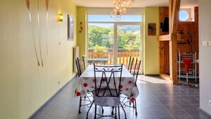 A dining area with a table and chairs, a view of greenery outside, and a kitchen area with a red chair. - Homerez - Comfortable Apartment with Garden (Saint-Dié-des-Vosges)