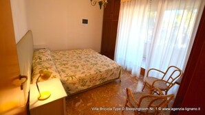 A bedroom with a bed, a chair, a lamp, a chandelier, and a window with curtains. - Lignano Exclusive With Pool (Lignano Sabbiadoro)