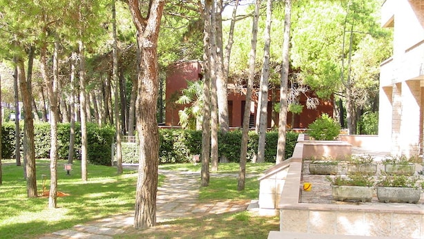 A courtyard with a stone pathway, trees, and a building in the background. - Lignano Exclusive With Pool (Lignano Sabbiadoro)
