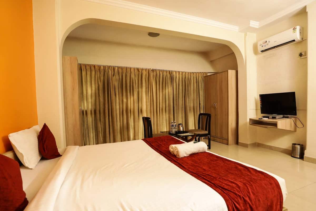A hotel room with a large bed, a TV, a desk with a chair, and an air conditioning unit.