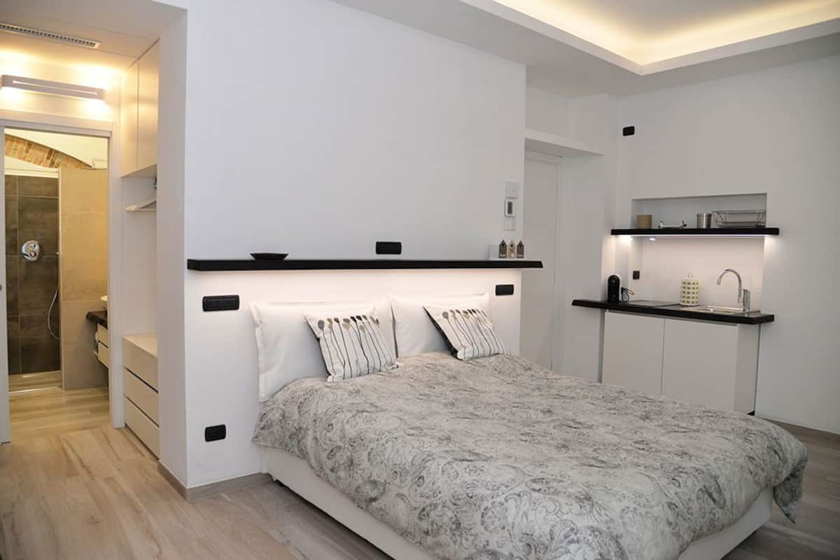 Ein modernes Schlafzimmer mit einem großen Bett, einer eingebauten Sitzbank, einer kleinen Küchenzeile und einem begehbaren Kleiderschrank.