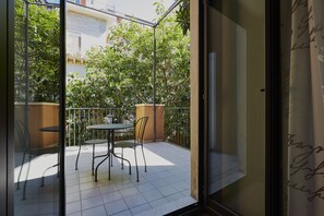 Un balcón con una mesa y sillas, una planta en maceta y vista a un árbol y un edificio.