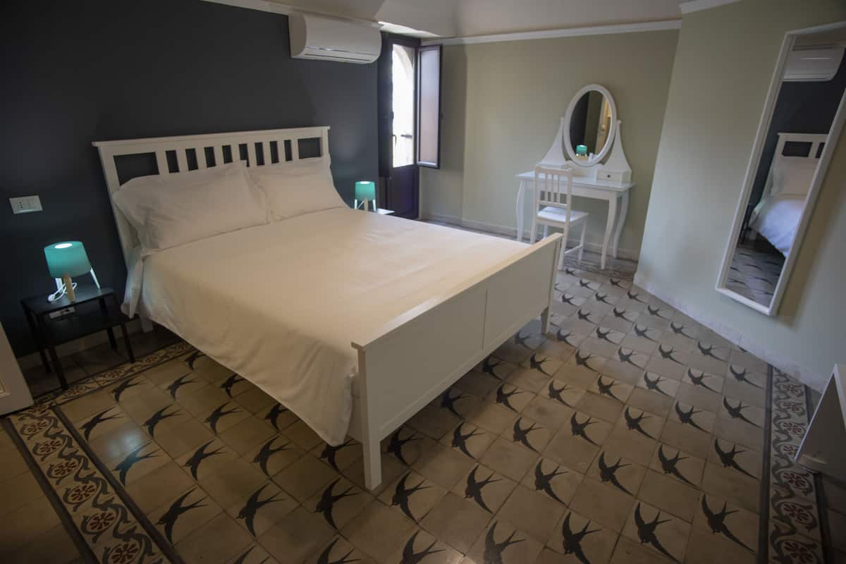 Een slaapkamer met een wit bed, een klein nachtkastje met een lamp, een witte kapstok met spiegel en een vloer met patroon.