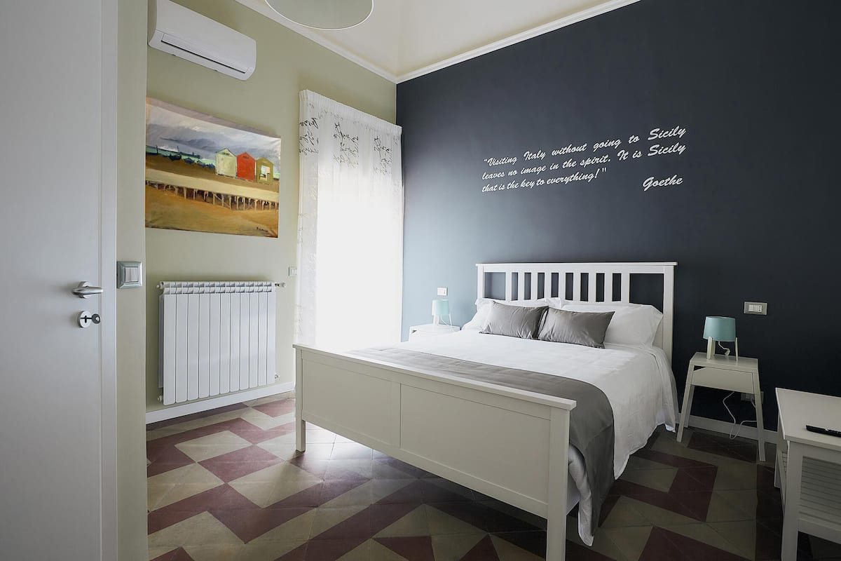 Een slaapkamer met een bed, nachtkastjes, een radiator, een schilderij en een citaat op de muur.