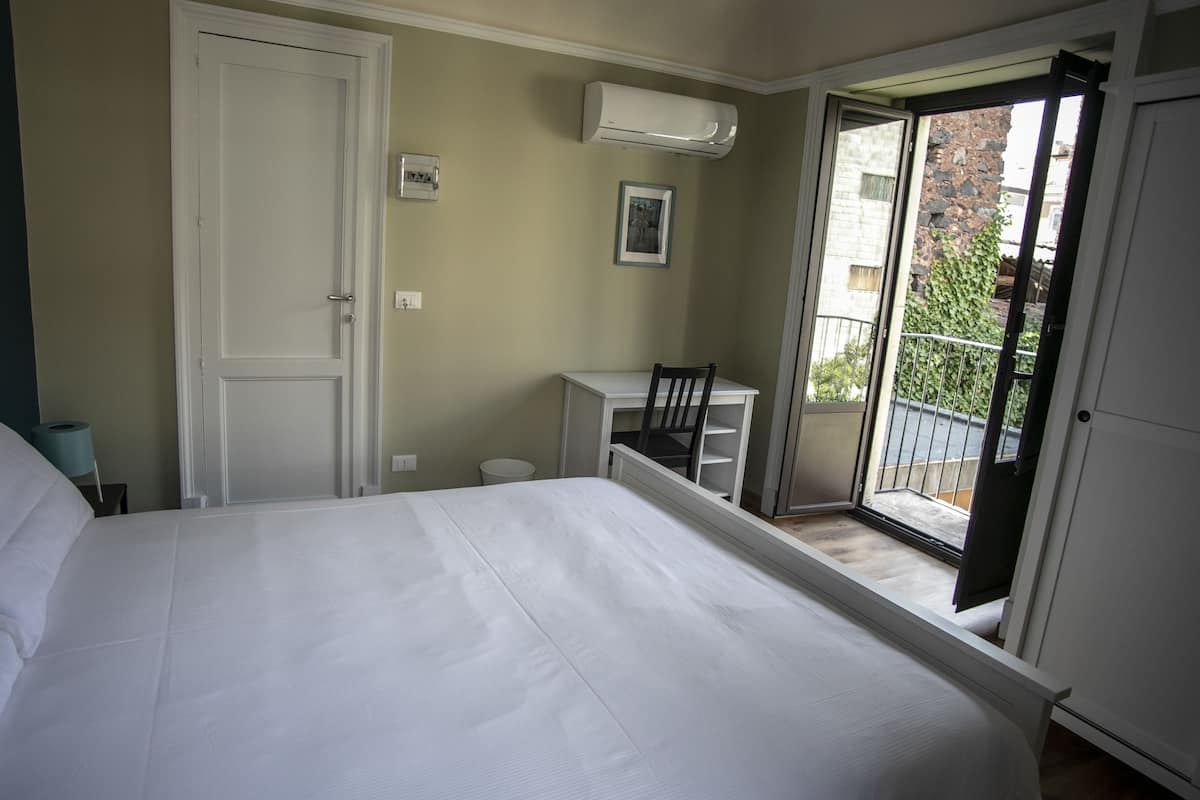 Een slaapkamer met een bed, een bureau, een stoel en een balkon met uitzicht op gebouwen en groen.