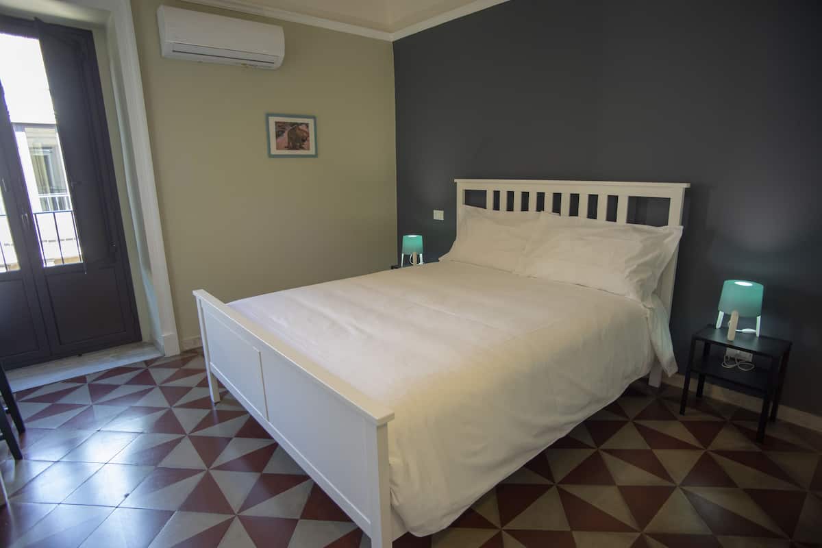 Een slaapkamer met een wit bed, een nachtkastje met een lamp, een deur en een raam.