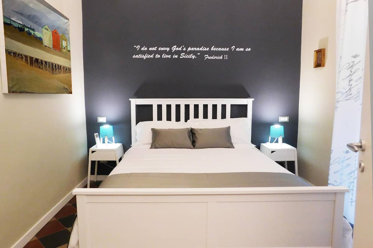 Een slaapkamer met een wit bed, twee nachtkastjes met lampen, en een citaat op de muur.