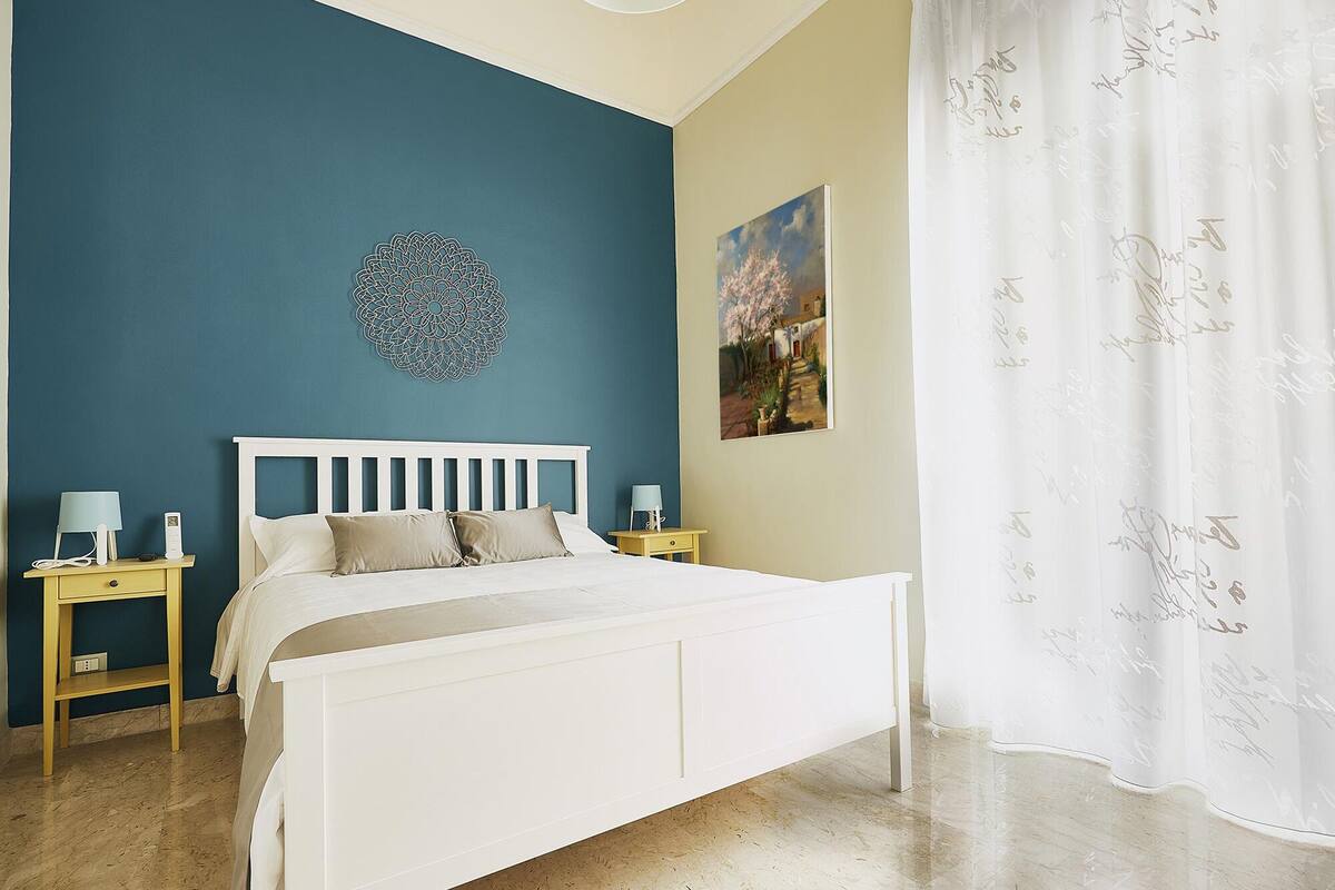 Een slaapkamer met een wit bed, een blauwe accentmuur, een decoratief wandobject, een schilderij en een wit gordijn met patroon.