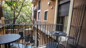 Un balcon avec deux tables rondes et quatre chaises, un arbre et un immeuble à la façade brune et aux fenêtres.