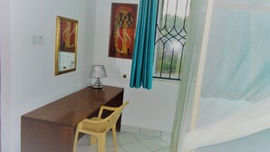 Un dormitorio con cama, escritorio, silla, ventana con cortinas y dos cuadros en la pared.