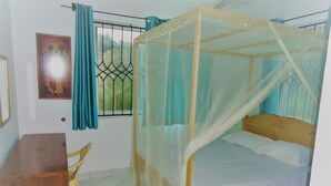 Una habitación con una cama bajo una mosquitera, un escritorio de madera, una silla, una ventana con barrotes y una puerta.
