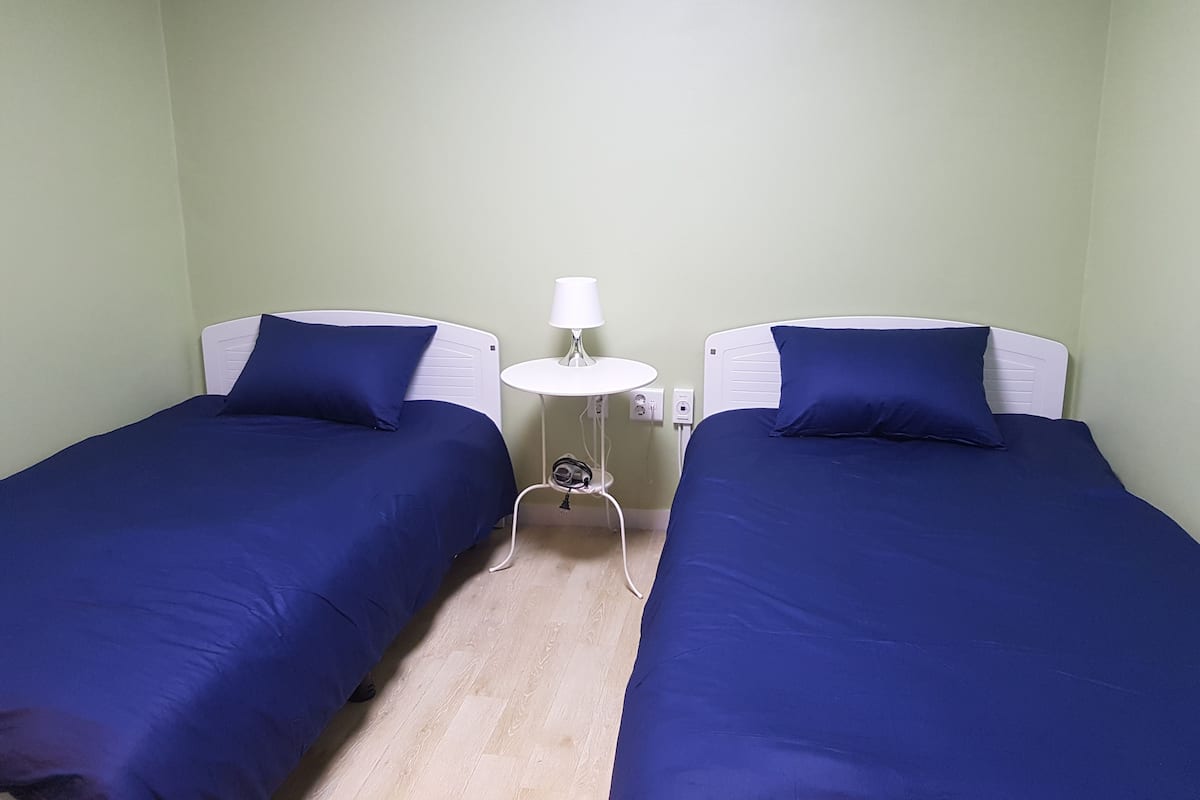 Ein Zimmer mit zwei Einzelbetten, jeweils mit weißem Kopfteil und blauer Bettdecke. Dazwischen steht ein kleiner weißer Tisch mit einer Lampe. Die Wände sind hellgrün gestrichen, und der Boden ist aus Holz.
