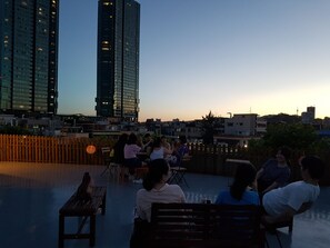 Terraza en la azotea con gente sentada en mesas, con vista a un paisaje urbano de edificios altos.
