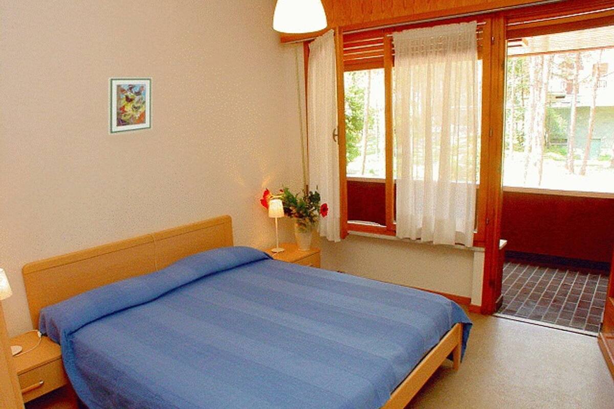Ein Schlafzimmer mit Bett, Nachttisch, Lampe, Fenster mit Vorhängen und einem kleinen Bild an der Wand.