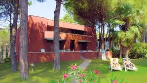 A modern building with a red facade, surrounded by lush greenery and palm trees. There are outdoor seating areas with white chairs.
