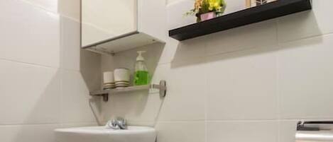 Un baño con lavamanos pedestal, inodoro y espejo sobre el lavamanos. Hay repisas sobre el lavamanos con artículos de aseo y un cuadro en la pared.