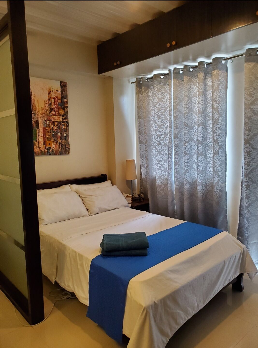 Una cama bien hecha con ropa de cama blanca y azul, un cuadro en la pared y una ventana con cortinas estampadas.