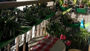 Un balcón con plantas en macetas, una mesa pequeña con mantel a cuadros y una silla.