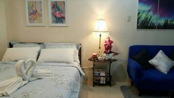 A bedroom with a bed, a blue sofa, a nightstand with a lamp, and wall art.