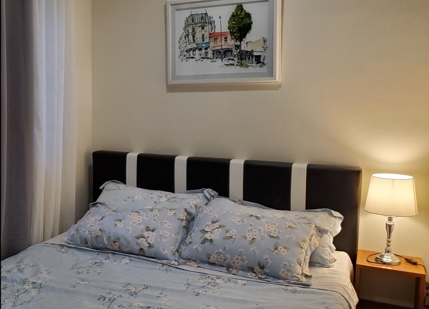 Una cama bien hecha con ropa de cama estampada con motivos florales, una obra de arte con un paisaje urbano enmarcado encima, y una lámpara de noche sobre una mesita de madera.