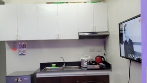 Una cocina compacta con armarios blancos, una encimera negra, un refrigerador pequeño, un hervidor y un televisor mostrando un programa.