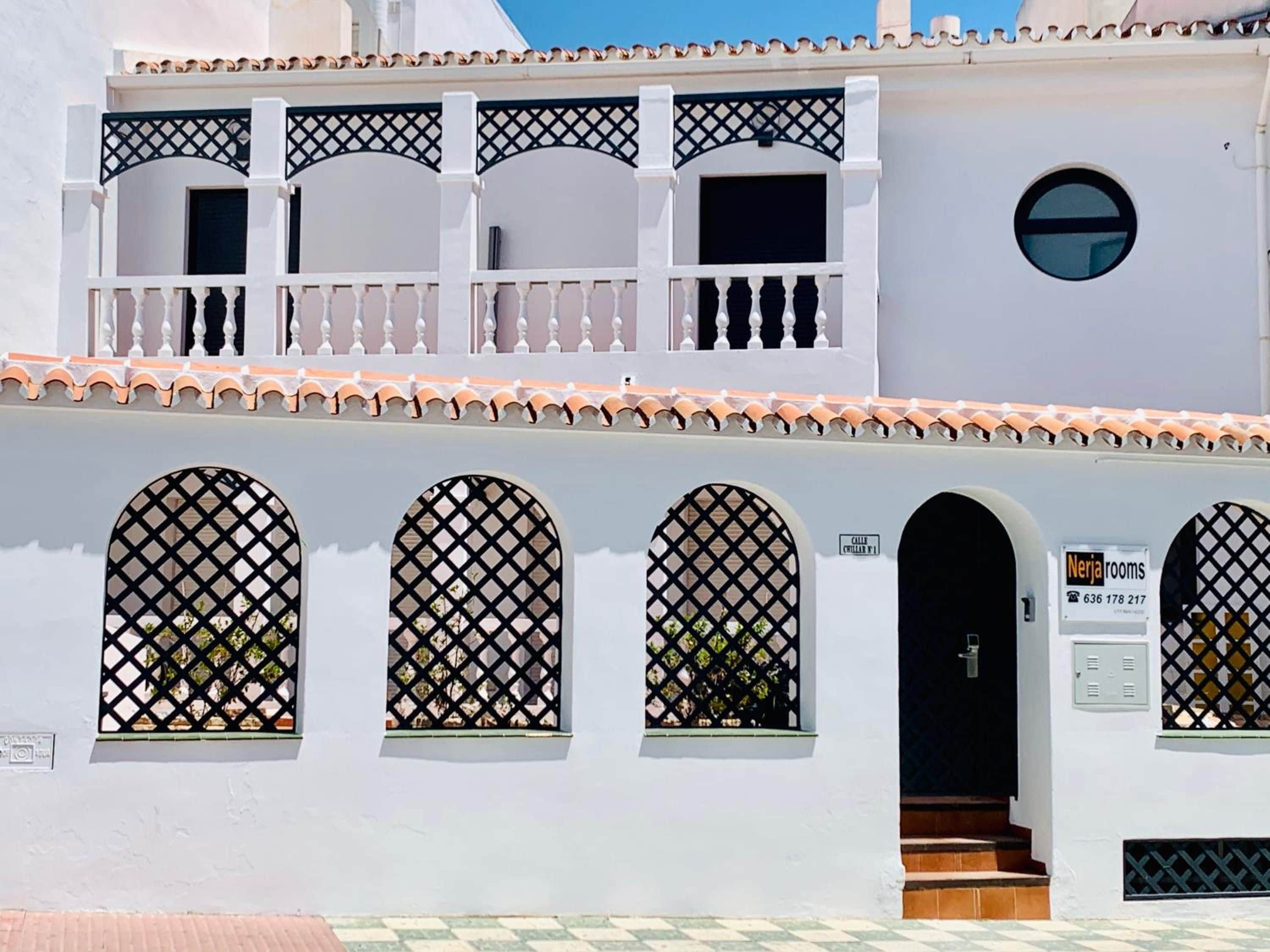Un bâtiment blanc doté d’un balcon, de trois fenêtres en arc surmontées de motifs en treillis et d’une porte sur laquelle est apposée une enseigne indiquant « Chambres à Nerja ».