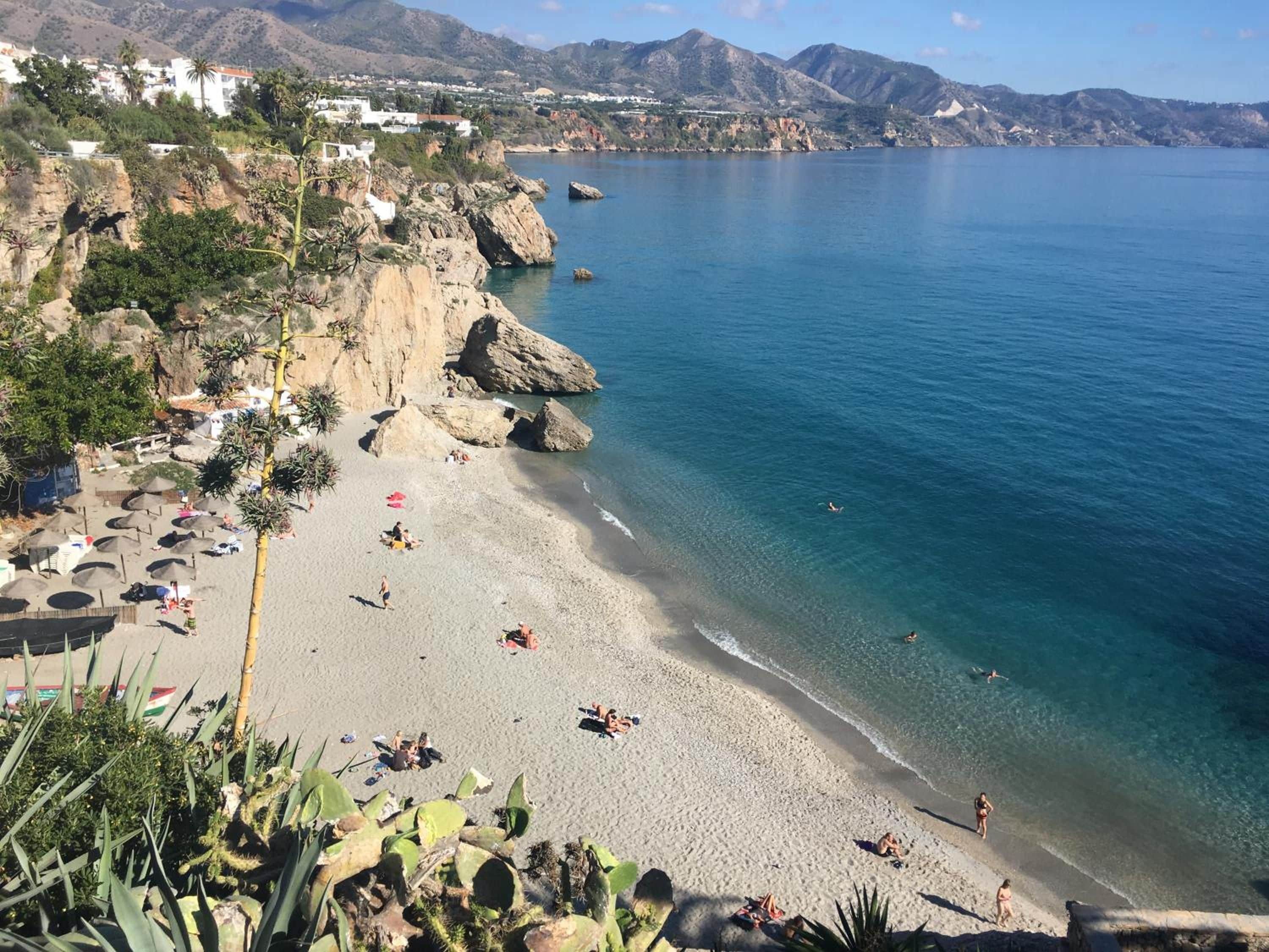 A coastal beach with clear waters, rocky cliffs, and a few people enjoying the sun.