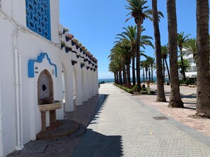 Une promenade en bord de mer bordée de bâtiments blancs, de fenêtres aux encadrements bleus et de palmiers.