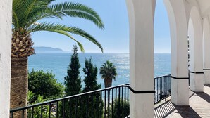 A balcony with a view of the sea, palm trees, and a white building with arches. - NerjaRooms (Nerja)