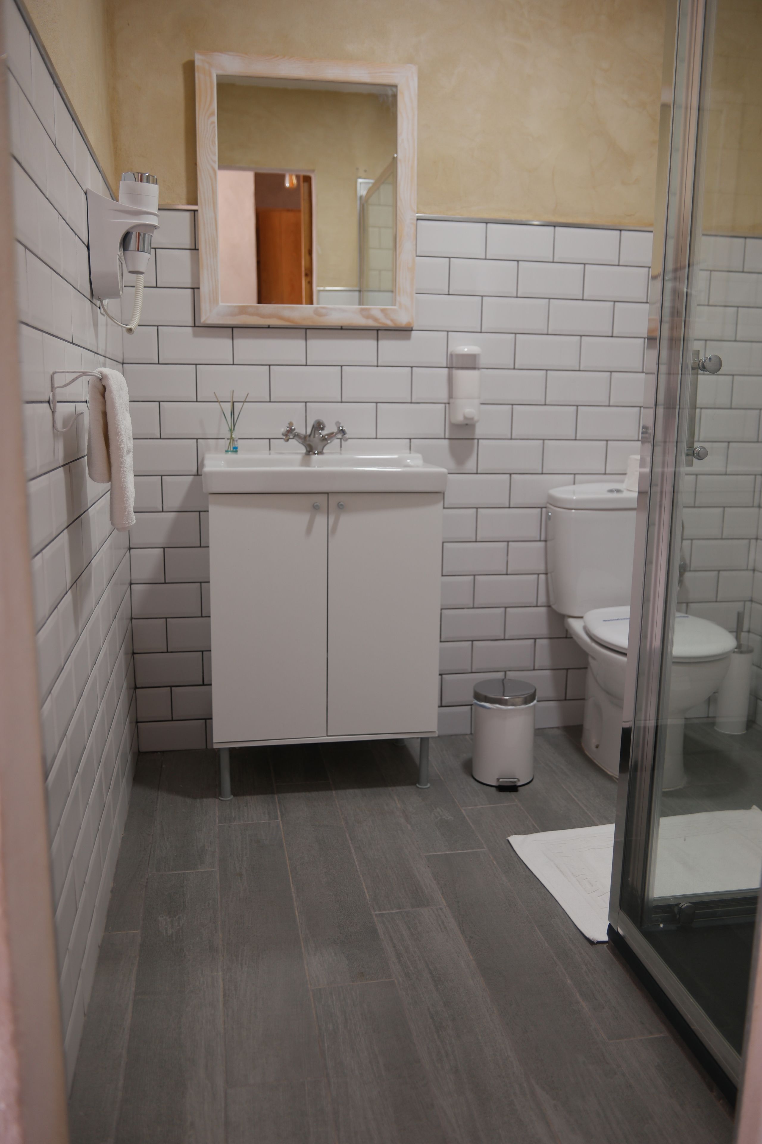 A bathroom with a white sink, mirror, and toilet, a trash bin, and a shower area.