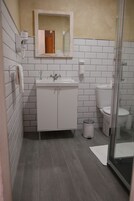 Un baño con lavamanos blanco, espejo, inodoro, un cubo de basura y zona de ducha.
